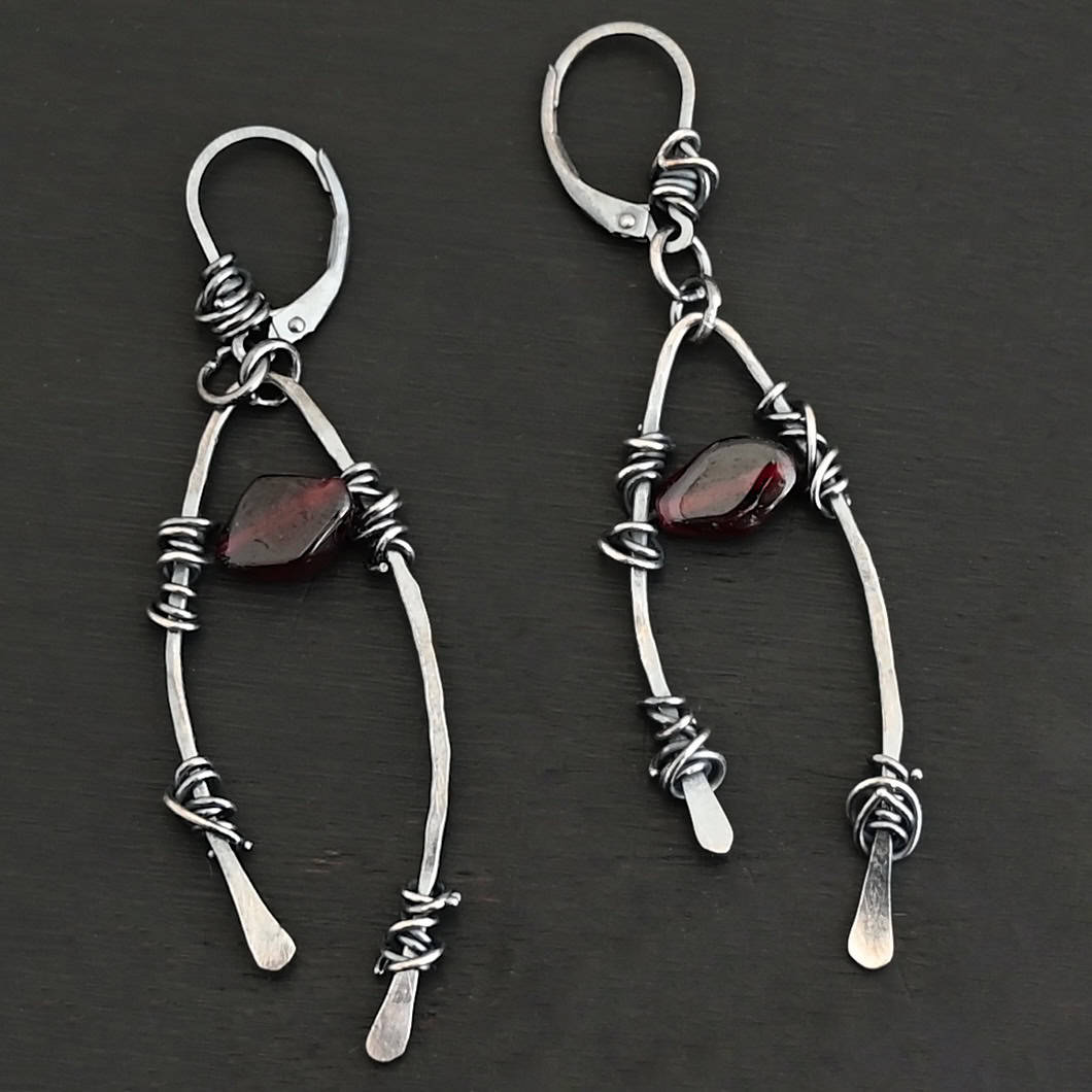 "Lanai" Earrings. There is an upside down "U" shape with silver wire wrapped around each side. There is a garnet bead in between the "U" shape for each earring.