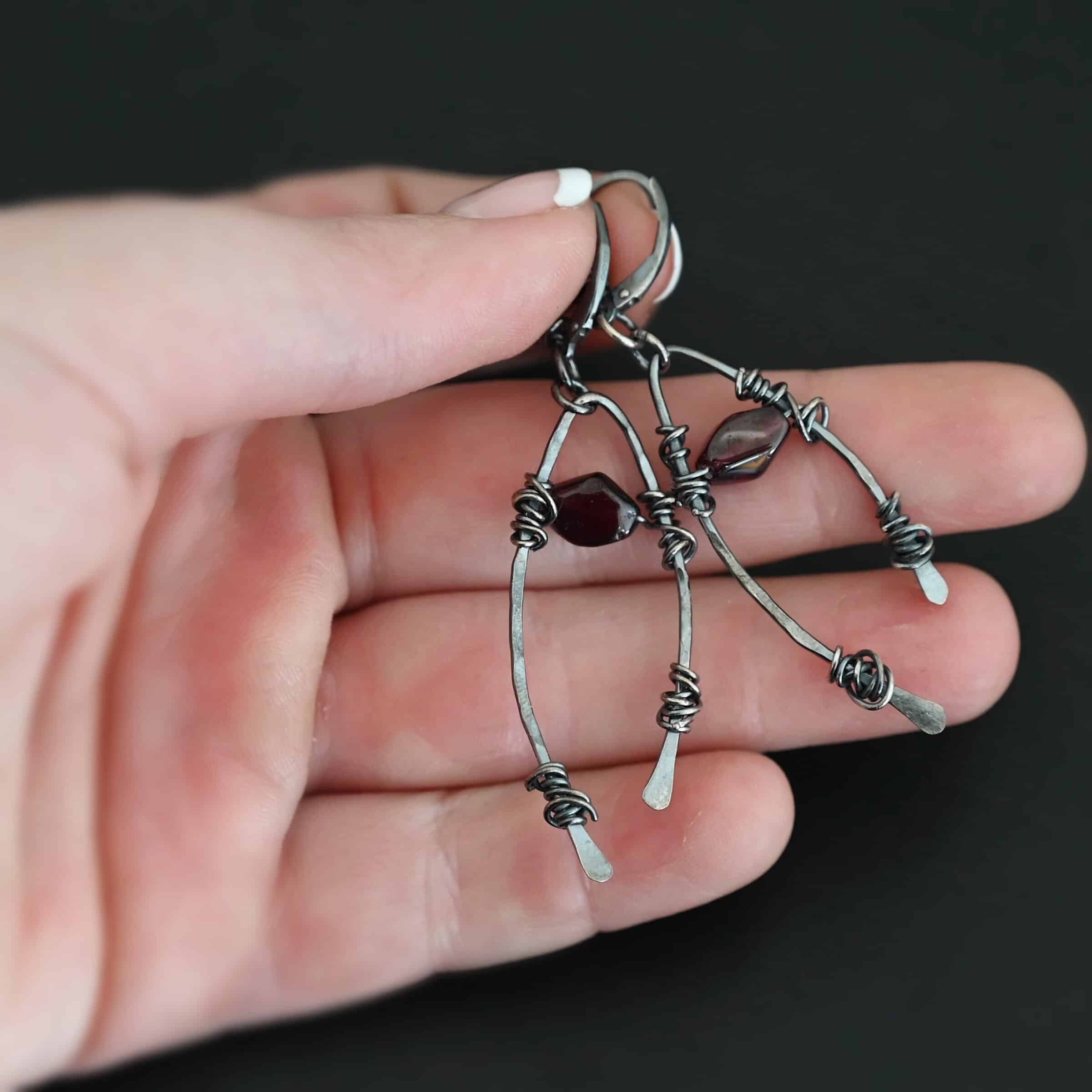 "Lanai" Earrings. There is an upside down "U" shape with silver wire wrapped around each side. There is a garnet bead in between the "U" shape for each earring. Earrings are held in the fingertips of a hand.