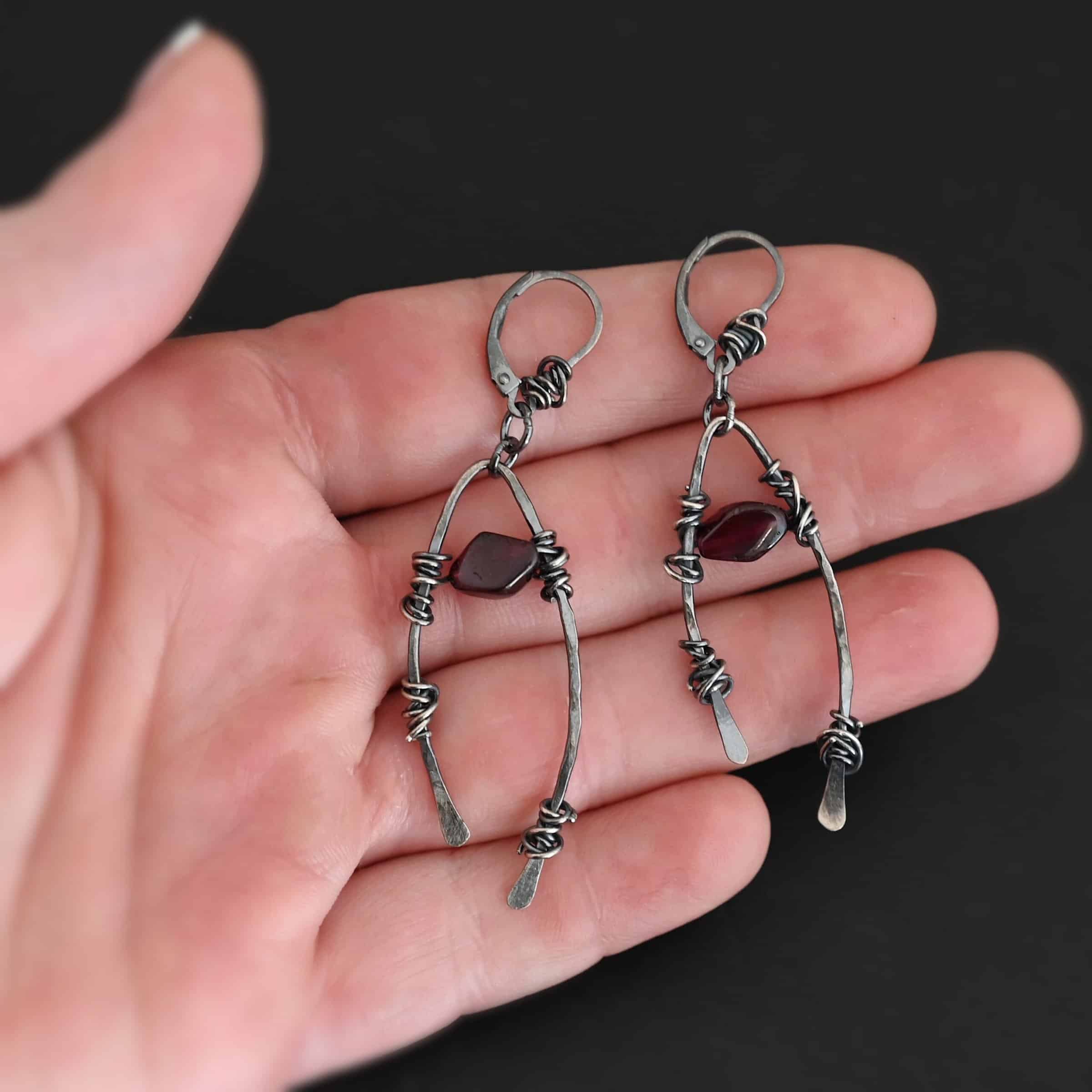 "Lanai" Earrings. There is an upside down "U" shape with silver wire wrapped around each side. There is a garnet bead in between the "U" shape for each earring. Earrings are held in the fingertips of a hand.