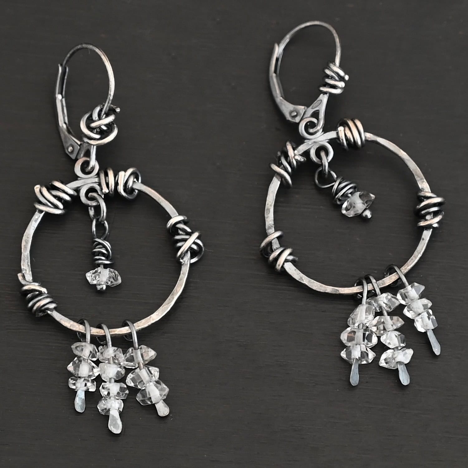 "Isla" Earrings. There is a hammered circle with silver wire wrapped around it in different parts. There are three small silver strands, each with three clear beads on it. There is a clear bead hanging in the center for each earring.