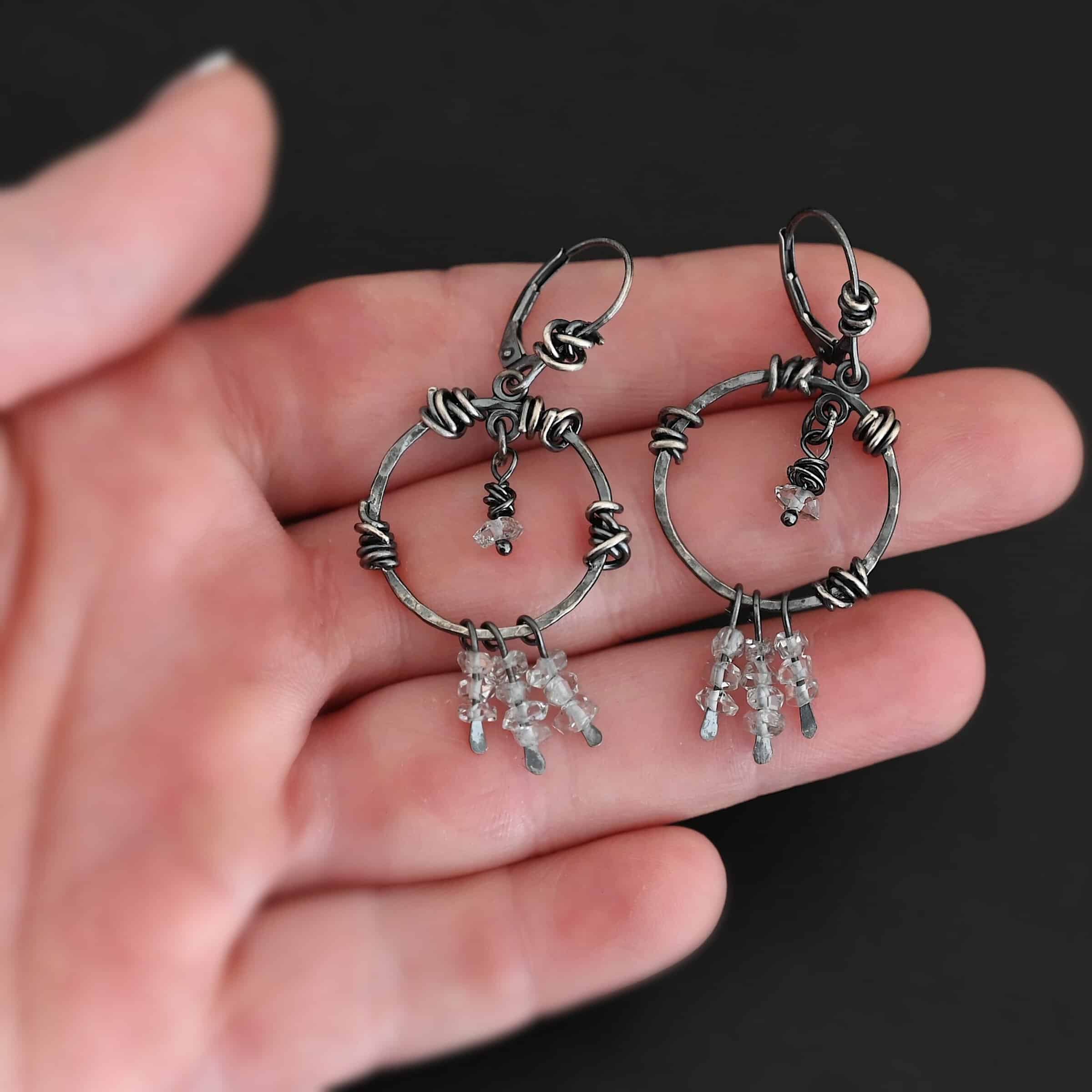 "Isla" Earrings. There is a hammered circle with silver wire wrapped around it in different parts. There are three small silver strands, each with three clear beads on it. There is a clear bead hanging in the center for each earring. Earrings are held in the fingertips of a hand.