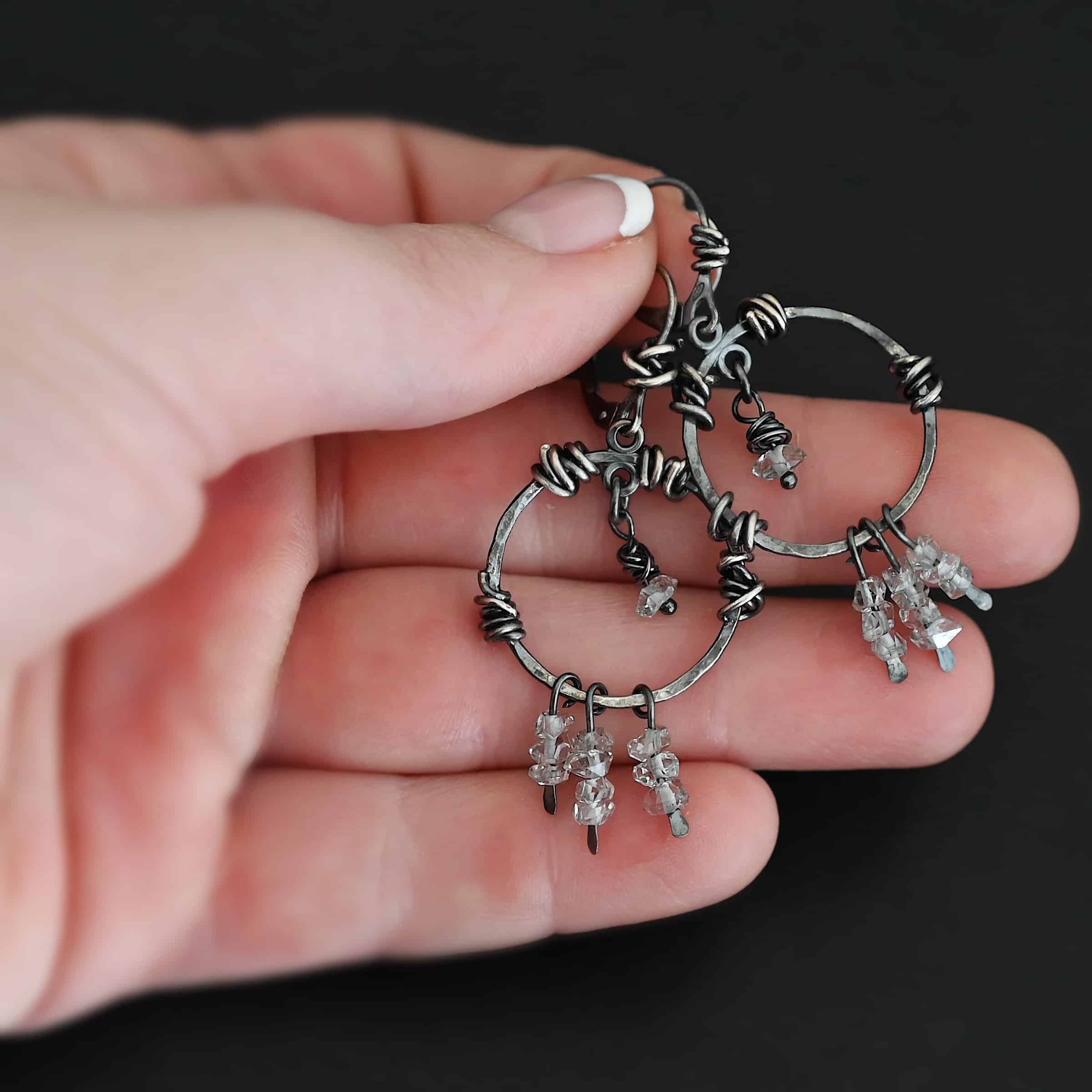 "Isla" Earrings. There is a hammered circle with silver wire wrapped around it in different parts. There are three small silver strands, each with three clear beads on it. There is a clear bead hanging in the center for each earring. Earrings are held in the fingertips of a hand.