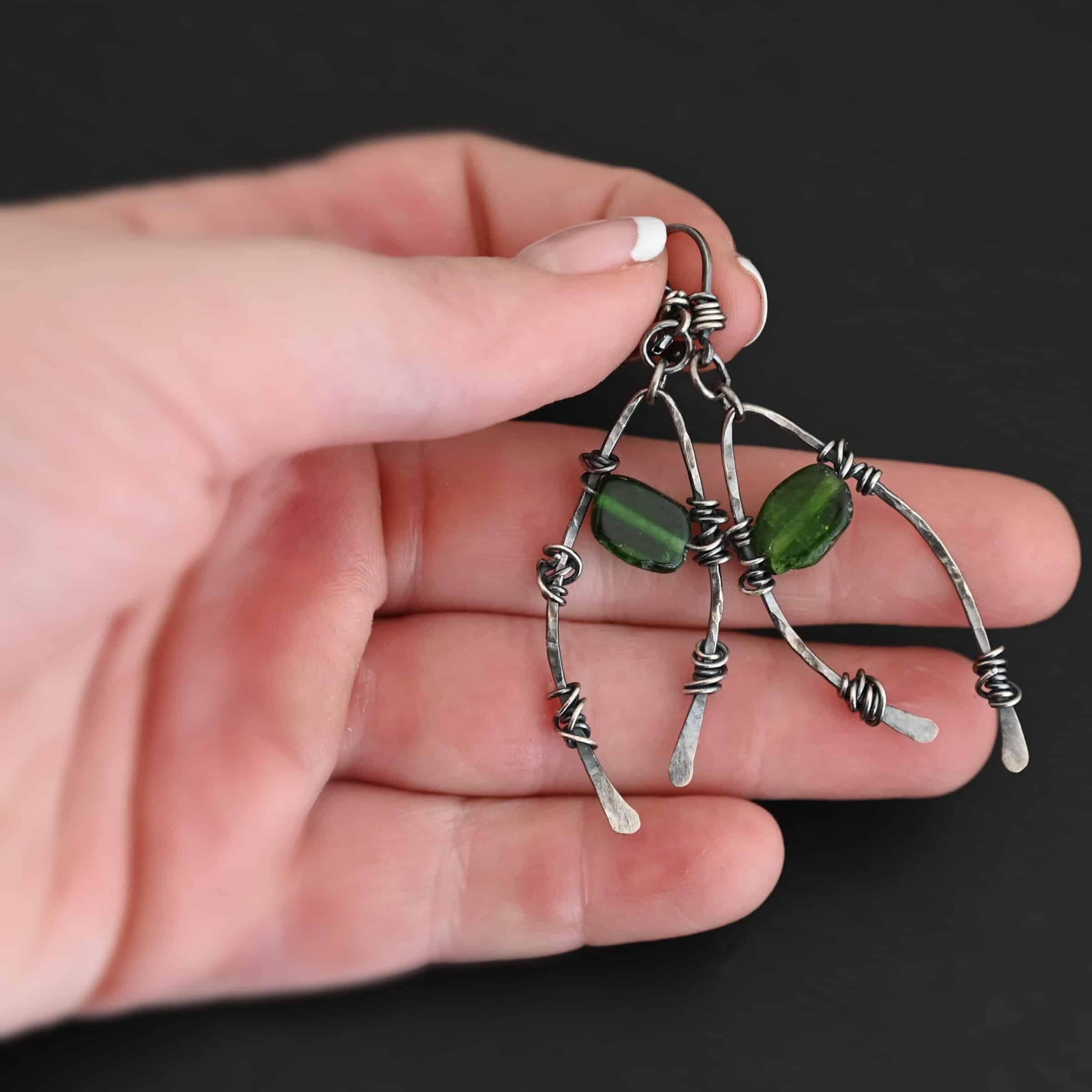 "Forest Glen" Earrings. There is an upside down "U" shape with silver wire wrapped around each side. There is a chrome diopside bead in between the "U" shape for each earring. Earrings are held in the fingertips of a hand.
