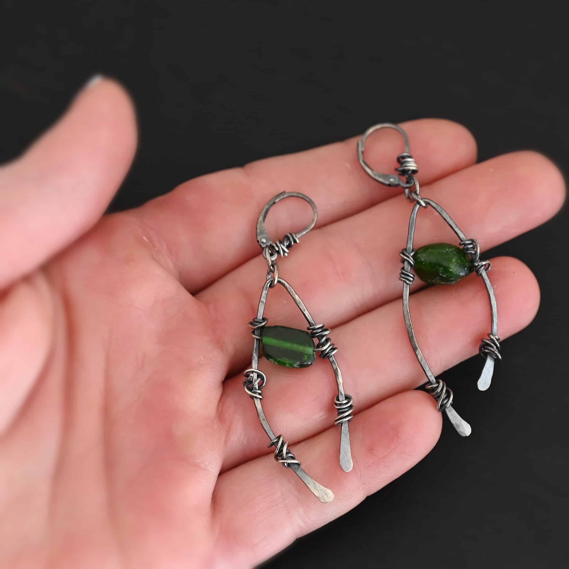 "Forest Glen" Earrings. There is an upside down "U" shape with silver wire wrapped around each side. There is a chrome diopside bead in between the "U" shape for each earring. Earrings are held in the fingertips of a hand.
