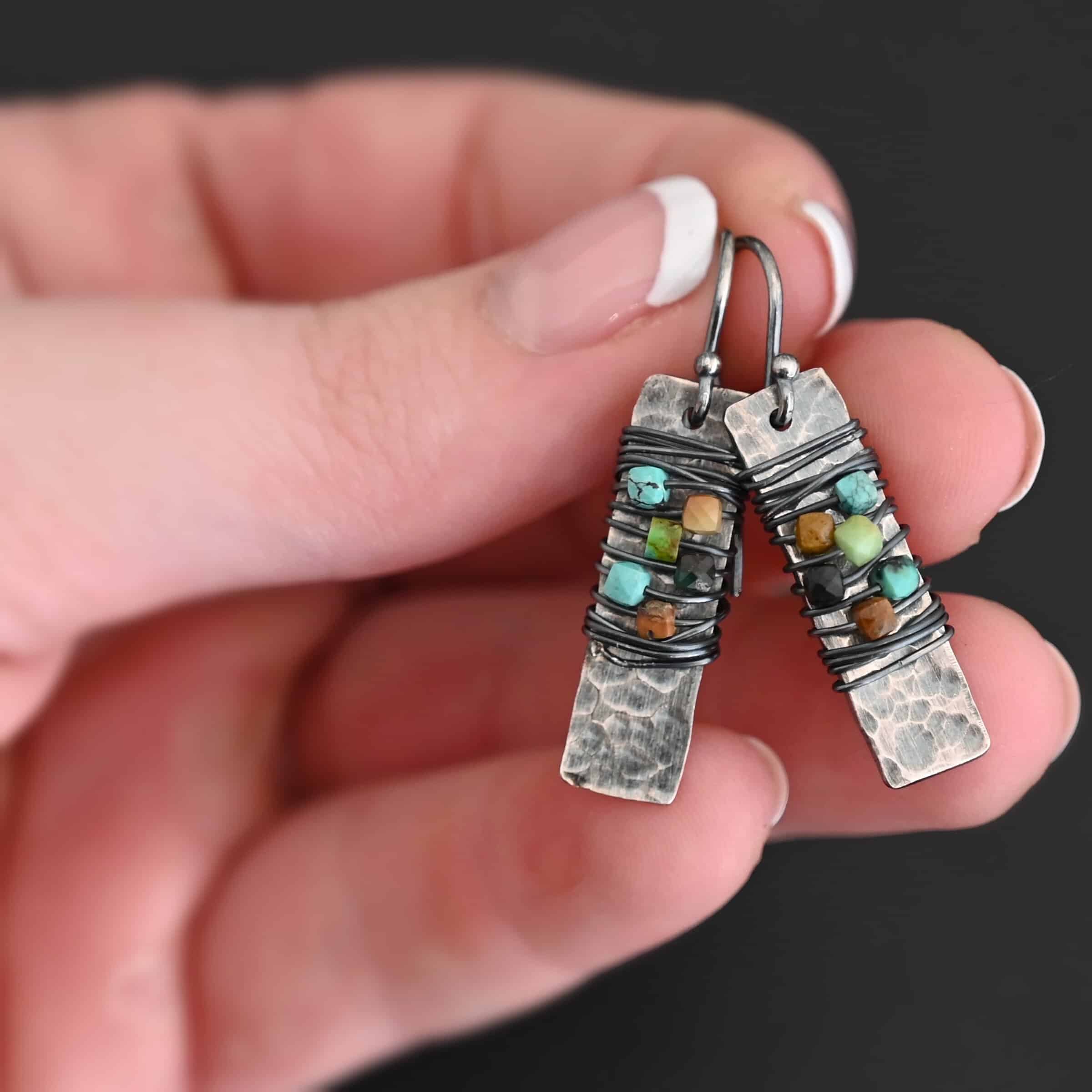 "Kea" Earrings. Each earring is a rectangular, vertical, hammered piece of silver. There is silver wire wrapped around the center of the earring, with five beads strung onto the wire. Earrings are held in the fingertips of a hand.