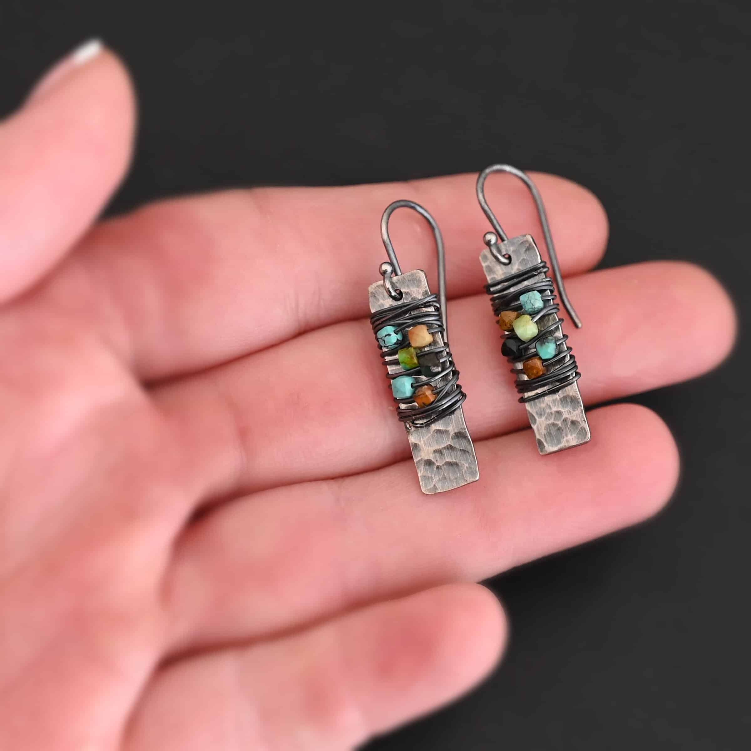"Kea" Earrings. Each earring is a rectangular, vertical, hammered piece of silver. There is silver wire wrapped around the center of the earring, with five beads strung onto the wire. Earrings are held in the fingertips of a hand.