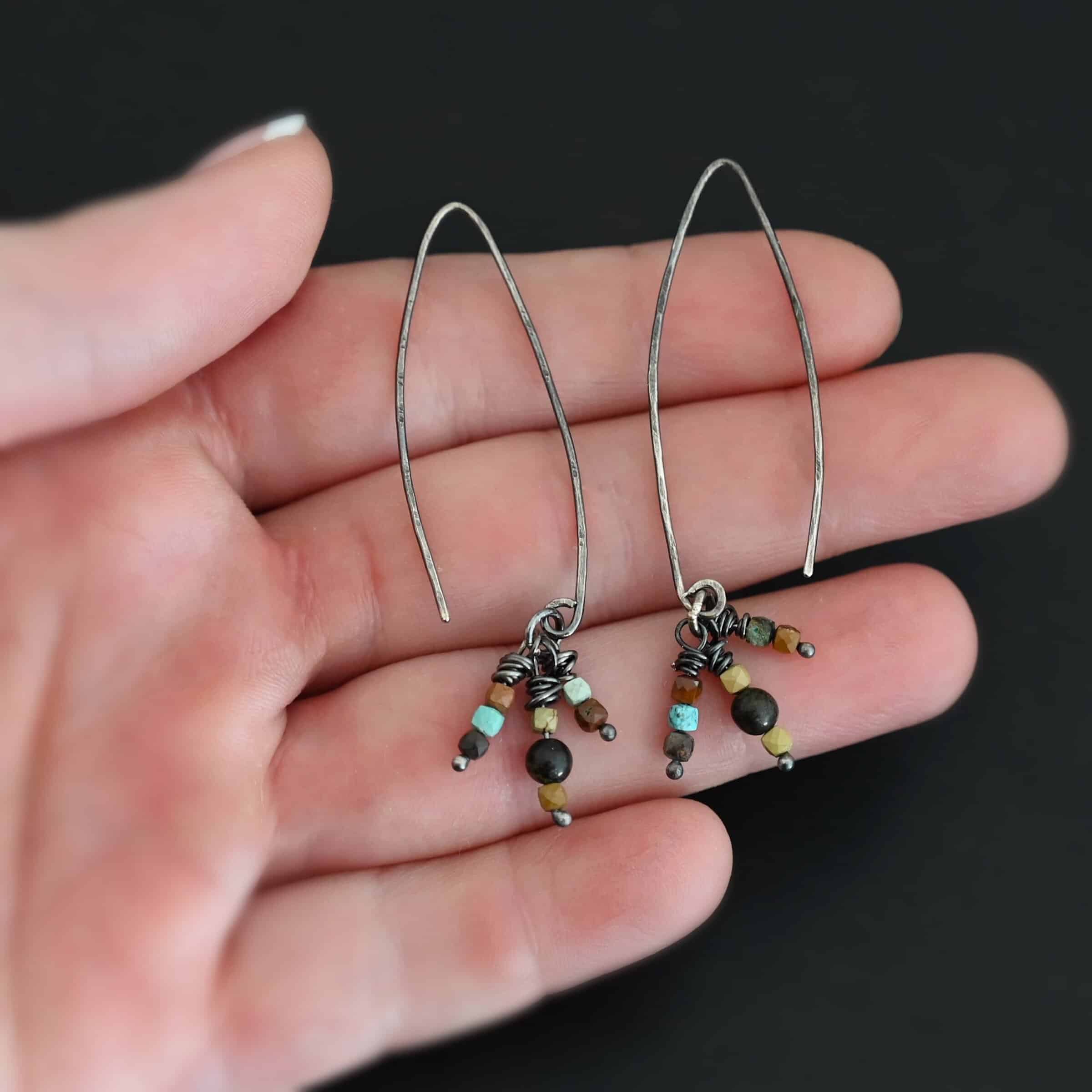 "Imber" Earrings. Each earring is an upside down "U" Shape. Each earring has three small strands of silver hanging down from it, each with three beads on it. Each strand has silver wrapped around the top. Earrings are held in the fingertips of a hand.