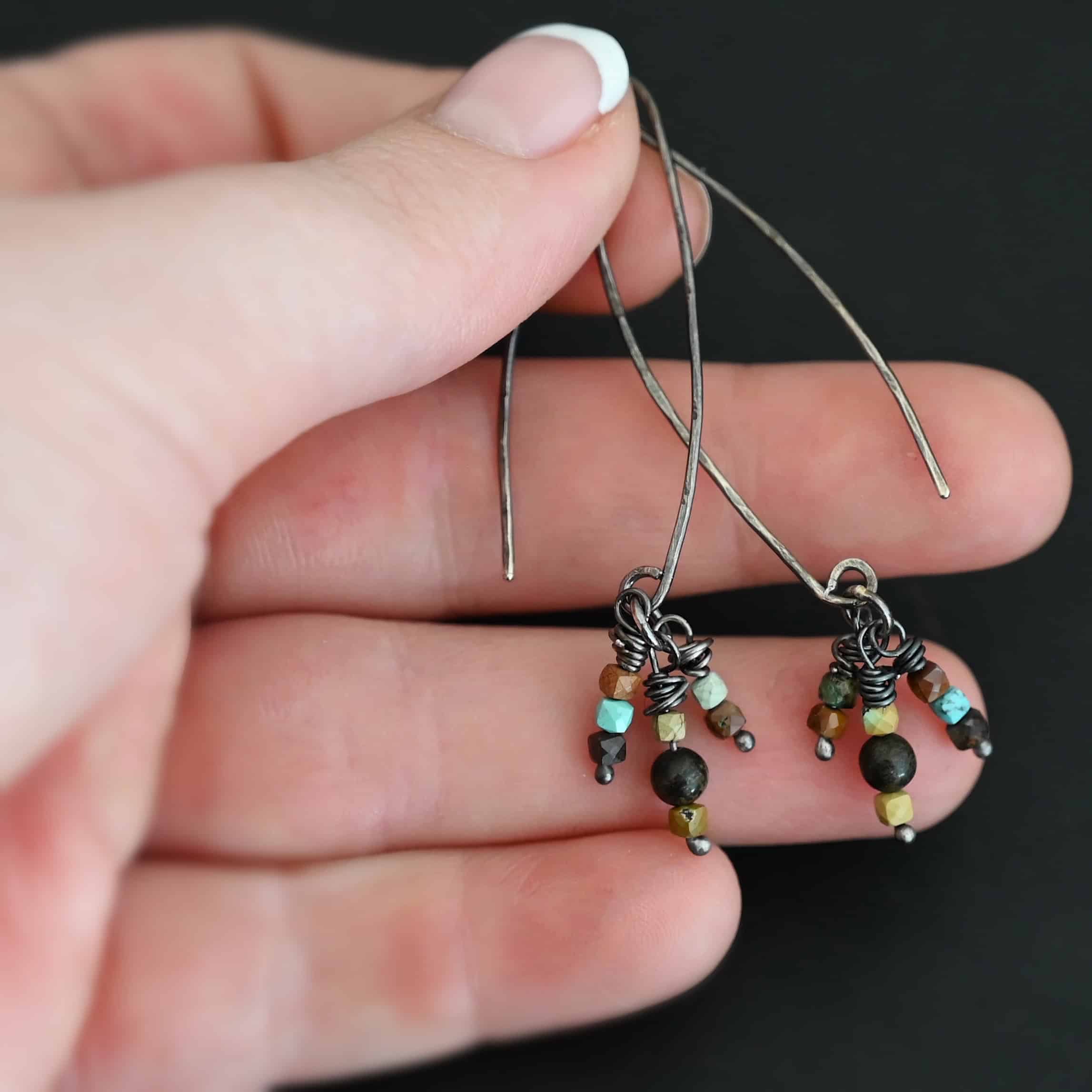 "Imber" Earrings. Each earring is an upside down "U" Shape. Each earring has three small strands of silver hanging down from it, each with three beads on it. Each strand has silver wrapped around the top. Earrings are held in the fingertips of a hand.