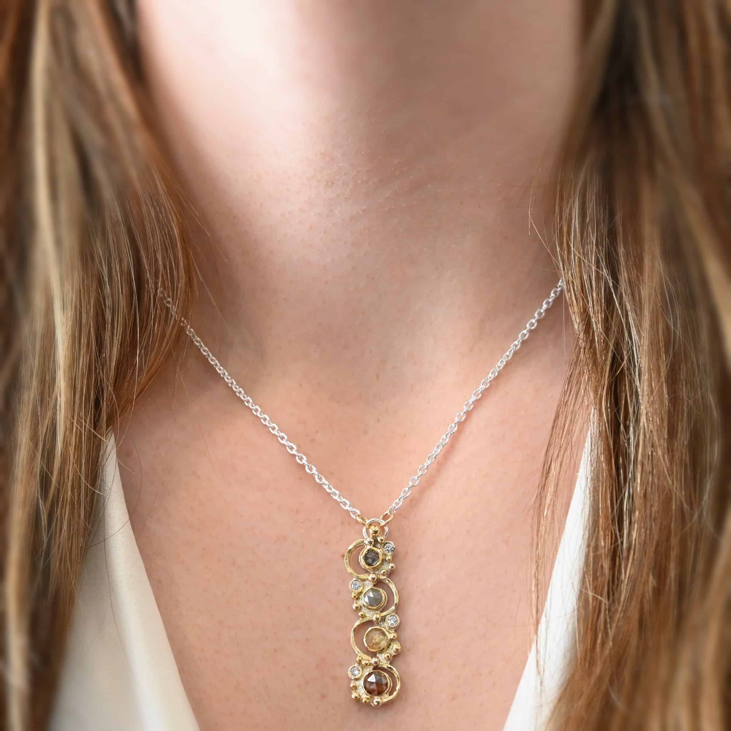 Photo of the "Music Of The Night" Necklace. Made out of sterling silver, gold, and diamonds. The top of the pendant, on the back, has a silver loop soldered to it with two gold jump rings that each end of the chain is attached to. There is gold ball detail that swirls back and forth all the way down the pendant. There is a line of hammered gold that also swirls around the pendant, but opposite of the gold balls, so they intertwine and intercept each other. There are eight diamonds on the pendant. Four of them are larger, and go in a straight line down the pendant. The other four are scattered around them. The gold line and gold balls twirl around each of the diamonds on opposite sides. Necklace is worn by a model at 17" long to demonstrate size.