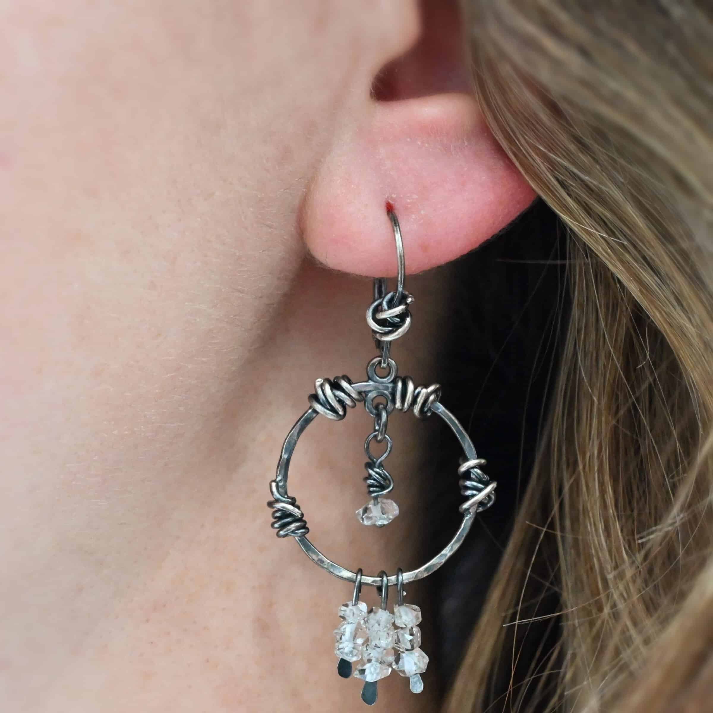 "Isla" Earring shown on a model. There is a hammered circle with silver wire wrapped around it in different parts. There are three small silver strands, each with three clear beads on it. There is a clear bead hanging in the center.