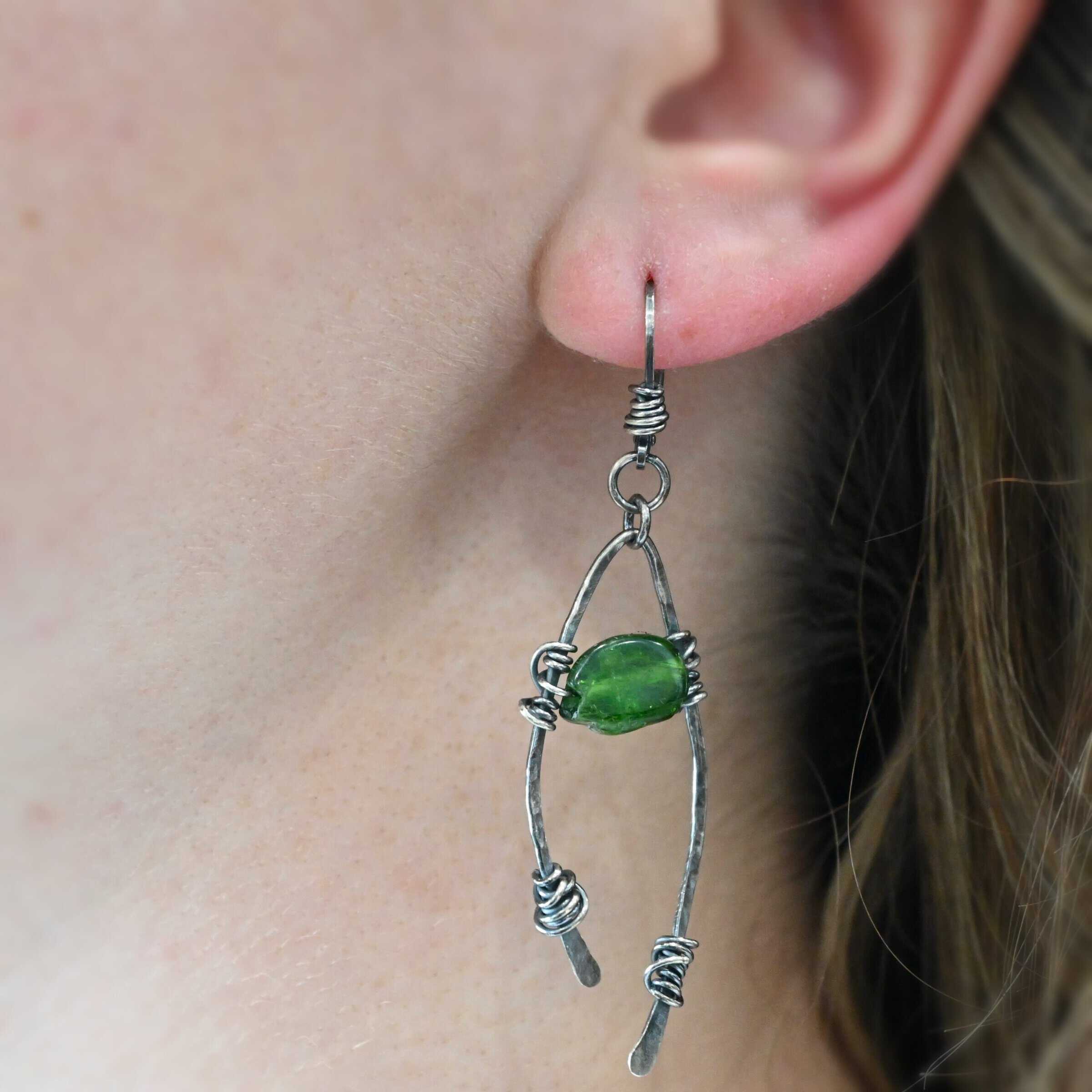 "Forest Glen" Earring shown on a model. There is an upside down "U" shape with silver wire wrapped around each side. There is a chrome diopside bead in between the "U" shape.