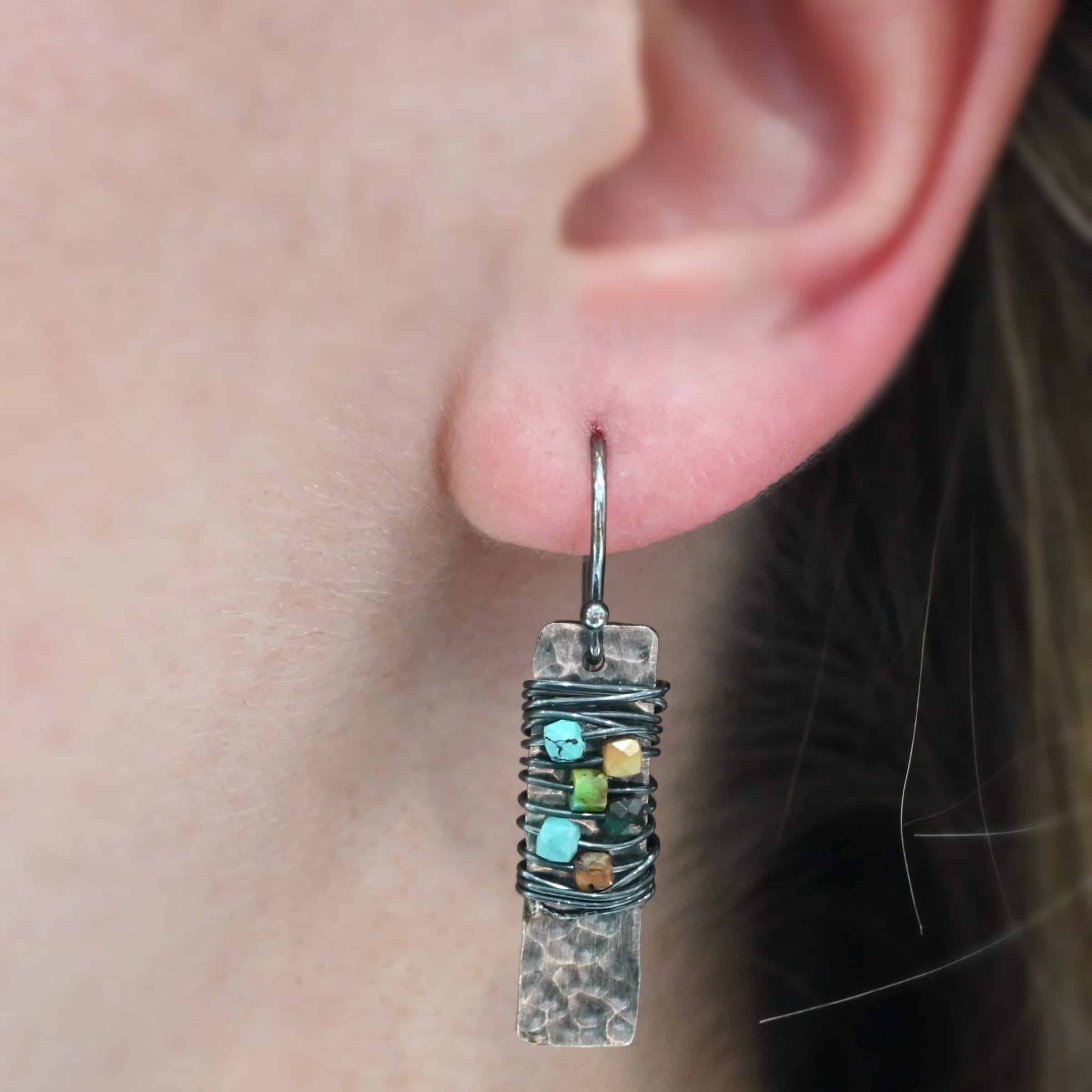 "Kea" Earring shown on a model. There is a rectangular, vertical, hammered piece of silver. There is silver wire wrapped around the center of the earring, with five beads strung onto the wire.