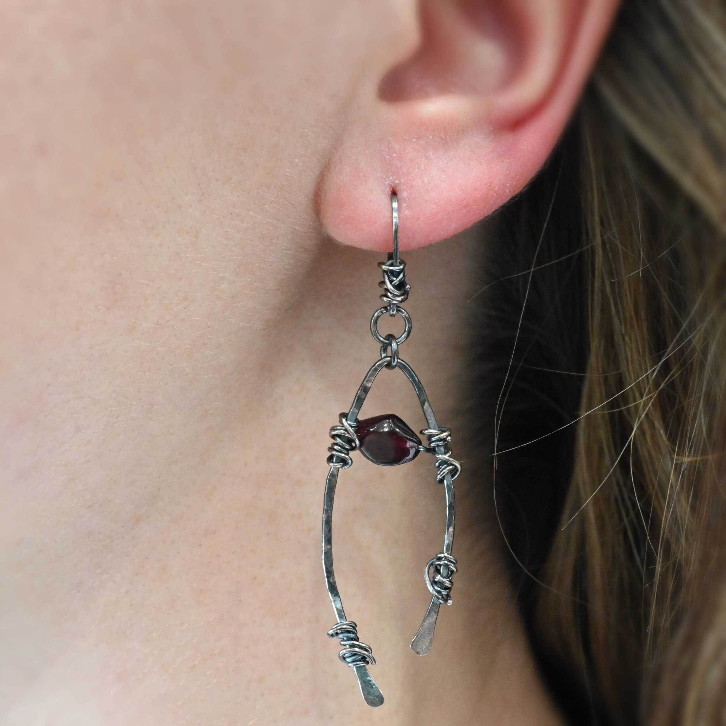 "Lanai" Earring shown on a model. There is an upside down "U" shape with silver wire wrapped around each side. There is a garnet bead in between the "U" shape.