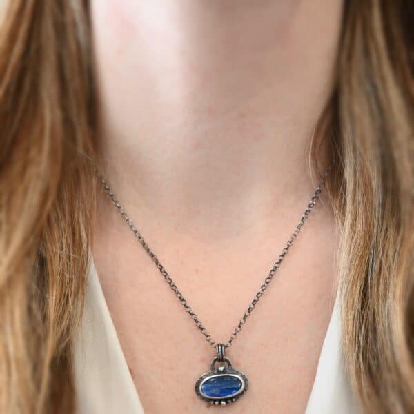 Photo of the "Caelum" Necklace. Made out of sterling silver and kyanite. There is an oval shaped deep blue kyanite set horizontally in the center. There is a ball of silver on the top of it in the center. There are five more balls of silver beneath it, that go in a straight line across the bottom. The rest of the silver around it is hammered. There is a loop soldered to the back with three jump rings attached to it. The chain for the necklace goes through the jump rings. Necklace is worn by a model.