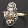 Photo of the "Garden Of Eden" Ring. Made out of sterling silver and diamonds. There is a ball of gold on the top and a small, clear, circular diamond beneath it. Then, a larger, darker green, circular diamond, and three balls of gold beneath that in a triangle shape. There are two layers of silver, both with hammered lines in them. The band is thin gold that is hammered.
