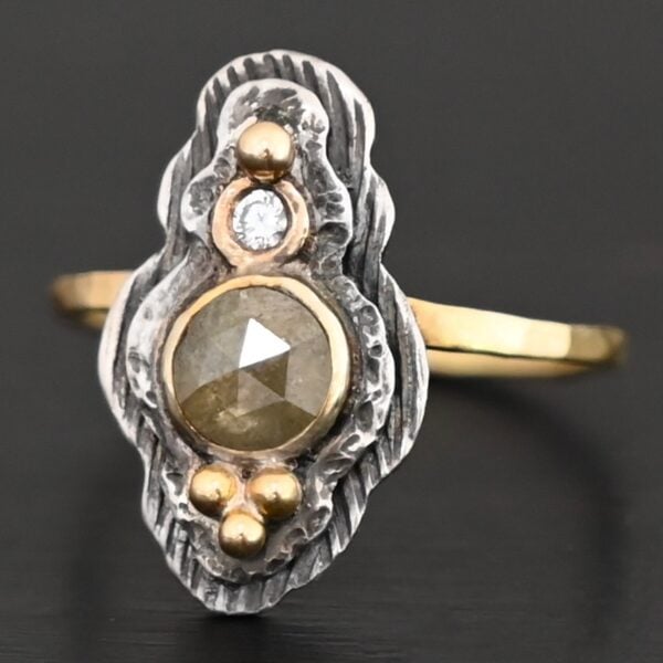 Photo of the "Garden Of Eden" Ring. Made out of sterling silver and diamonds. There is a ball of gold on the top and a small, clear, circular diamond beneath it. Then, a larger, darker green, circular diamond, and three balls of gold beneath that in a triangle shape. There are two layers of silver, both with hammered lines in them. The band is thin gold that is hammered.