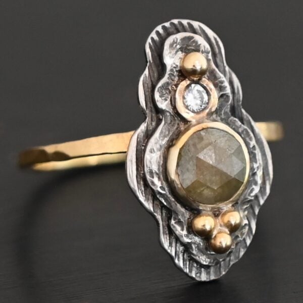Photo of the "Garden Of Eden" Ring. Made out of sterling silver and diamonds. There is a ball of gold on the top and a small, clear, circular diamond beneath it. Then, a larger, darker green, circular diamond, and three balls of gold beneath that in a triangle shape. There are two layers of silver, both with hammered lines in them. The band is thin gold that is hammered.