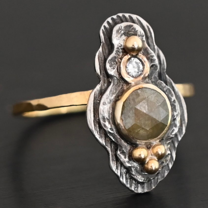 Garden Of Eden Ring New Photo of the "Garden Of Eden" Ring. Made out of sterling silver and diamonds. There is a ball of gold on the top and a small, clear, circular diamond beneath it. Then, a larger, darker green, circular diamond, and three balls of gold beneath that in a triangle shape. There are two layers of silver, both with hammered lines in them. The band is thin gold that is hammered.