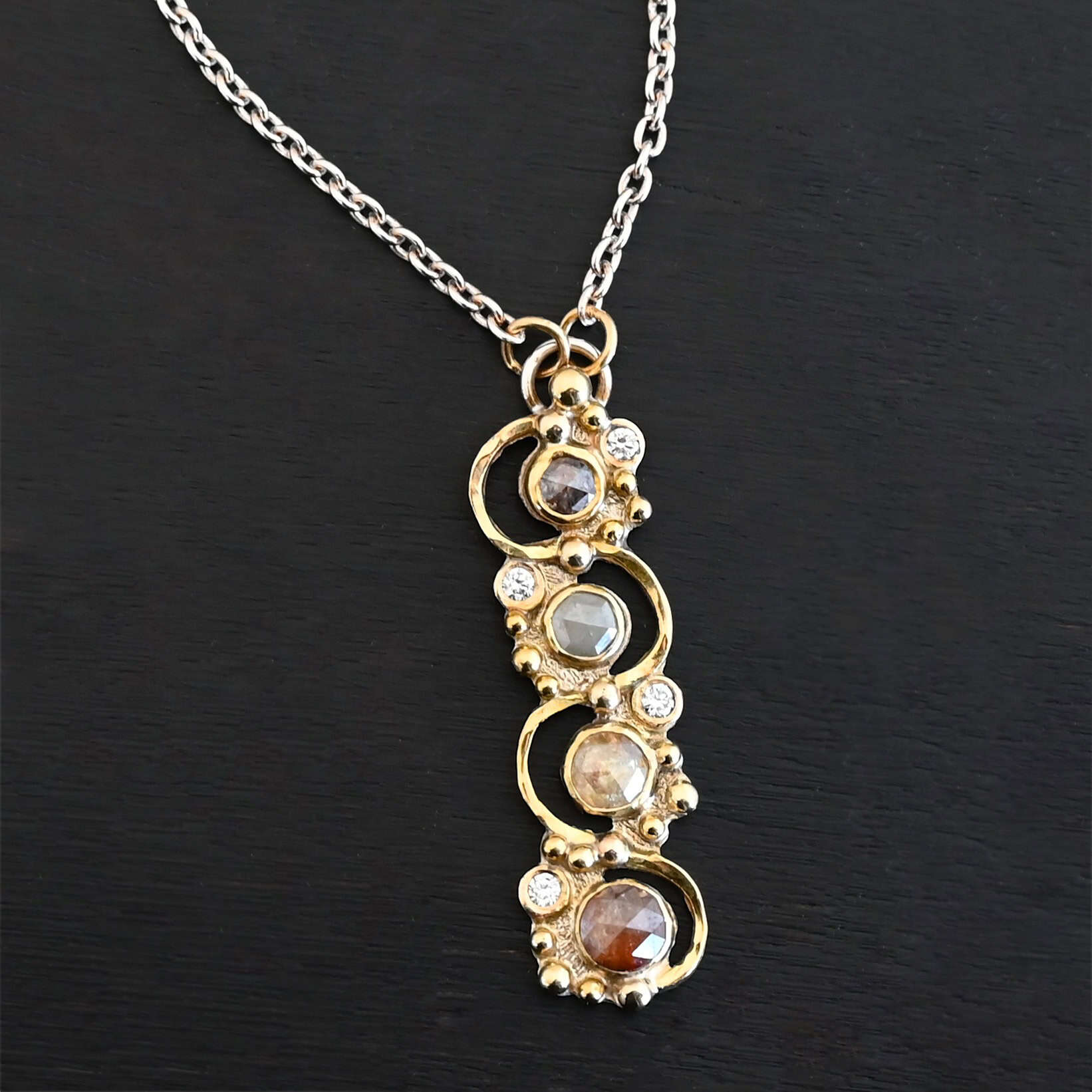 Photo of the "Music Of The Night" Necklace. Made out of sterling silver, gold, and diamonds. The top of the pendant, on the back, has a silver loop soldered to it with two gold jump rings that each end of the chain is attached to. There is gold ball detail that swirls back and forth all the way down the pendant. There is a line of hammered gold that also swirls around the pendant, but opposite of the gold balls, so they intertwine and intercept each other. There are eight diamonds on the pendant. Four of them are larger, and go in a straight line down the pendant. The other four are scattered around them. The gold line and gold balls twirl around each of the diamonds on opposite sides. Necklace is lying against a dark background.