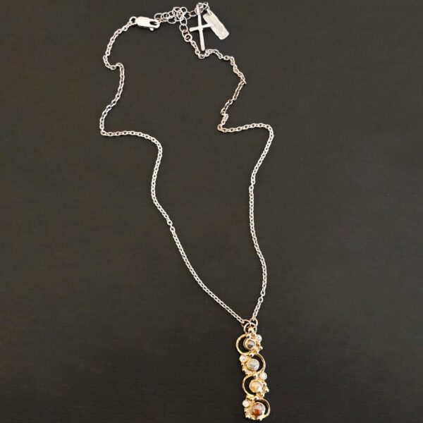Photo of the "Music Of The Night" Necklace. Made out of sterling silver, gold, and diamonds. The top of the pendant, on the back, has a silver loop soldered to it with two gold jump rings that each end of the chain is attached to. There is gold ball detail that swirls back and forth all the way down the pendant. There is a line of hammered gold that also swirls around the pendant, but opposite of the gold balls, so they intertwine and intercept each other. There are eight diamonds on the pendant. Four of them are larger, and go in a straight line down the pendant. The other four are scattered around them. The gold line and gold balls twirl around each of the diamonds on opposite sides. Necklace is lying against a dark background.