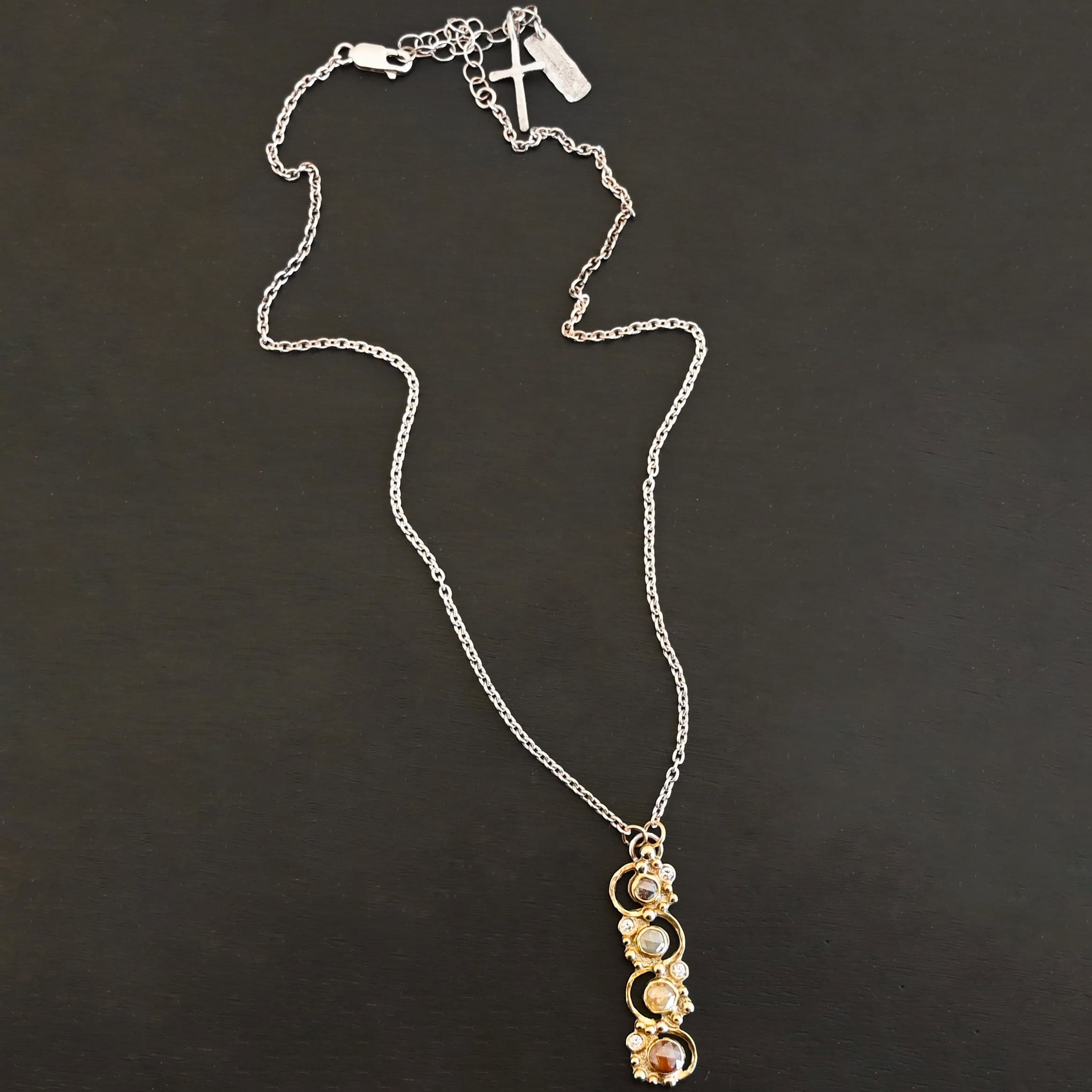 Photo of the "Music Of The Night" Necklace. Made out of sterling silver, gold, and diamonds. The top of the pendant, on the back, has a silver loop soldered to it with two gold jump rings that each end of the chain is attached to. There is gold ball detail that swirls back and forth all the way down the pendant. There is a line of hammered gold that also swirls around the pendant, but opposite of the gold balls, so they intertwine and intercept each other. There are eight diamonds on the pendant. Four of them are larger, and go in a straight line down the pendant. The other four are scattered around them. The gold line and gold balls twirl around each of the diamonds on opposite sides. Necklace is lying against a dark background.