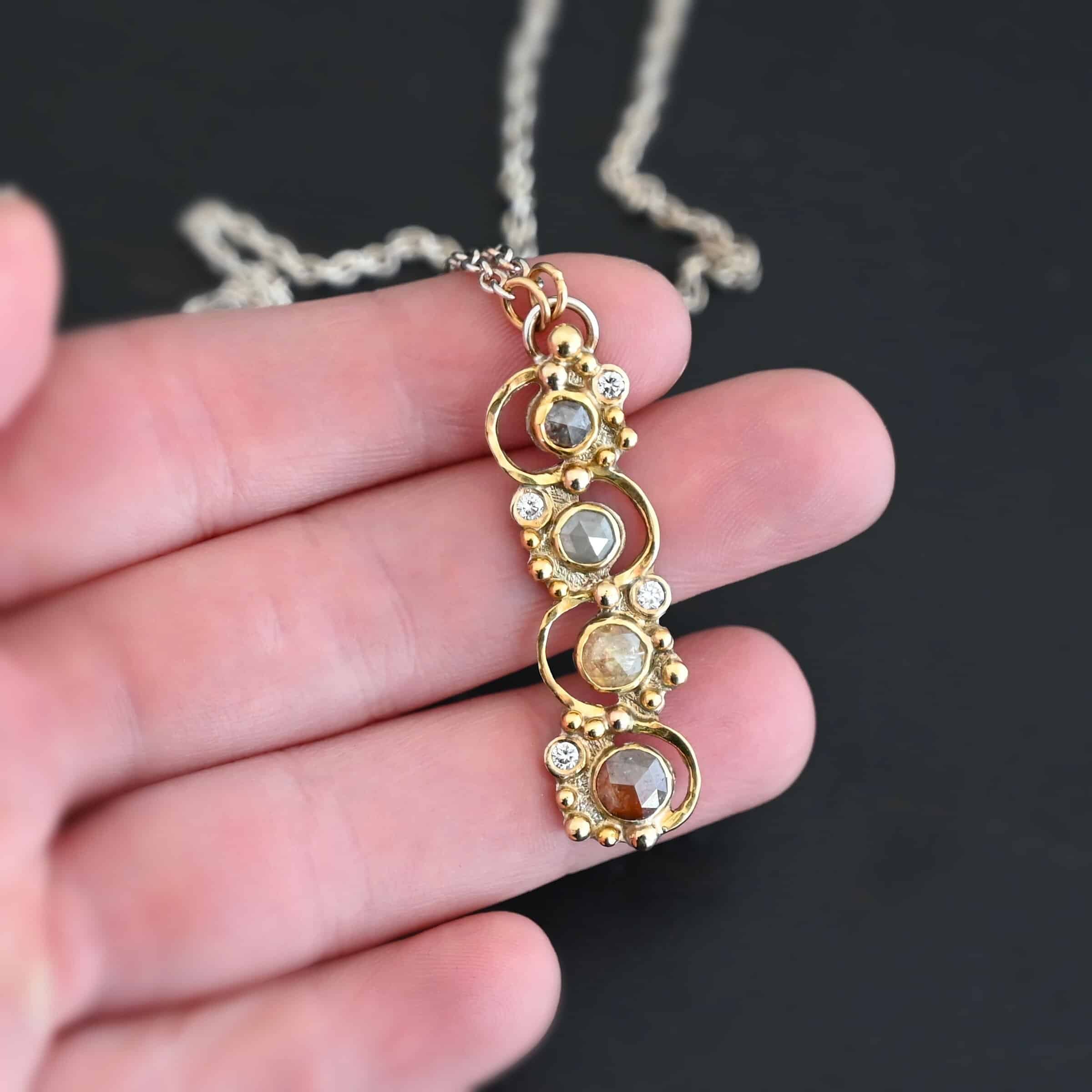 Photo of the "Music Of The Night" Necklace. Made out of sterling silver, gold, and diamonds. The top of the pendant, on the back, has a silver loop soldered to it with two gold jump rings that each end of the chain is attached to. There is gold ball detail that swirls back and forth all the way down the pendant. There is a line of hammered gold that also swirls around the pendant, but opposite of the gold balls, so they intertwine and intercept each other. There are eight diamonds on the pendant. Four of them are larger, and go in a straight line down the pendant. The other four are scattered around them. The gold line and gold balls twirl around each of the diamonds on opposite sides. Necklace is lying against the fingertips of a hand.