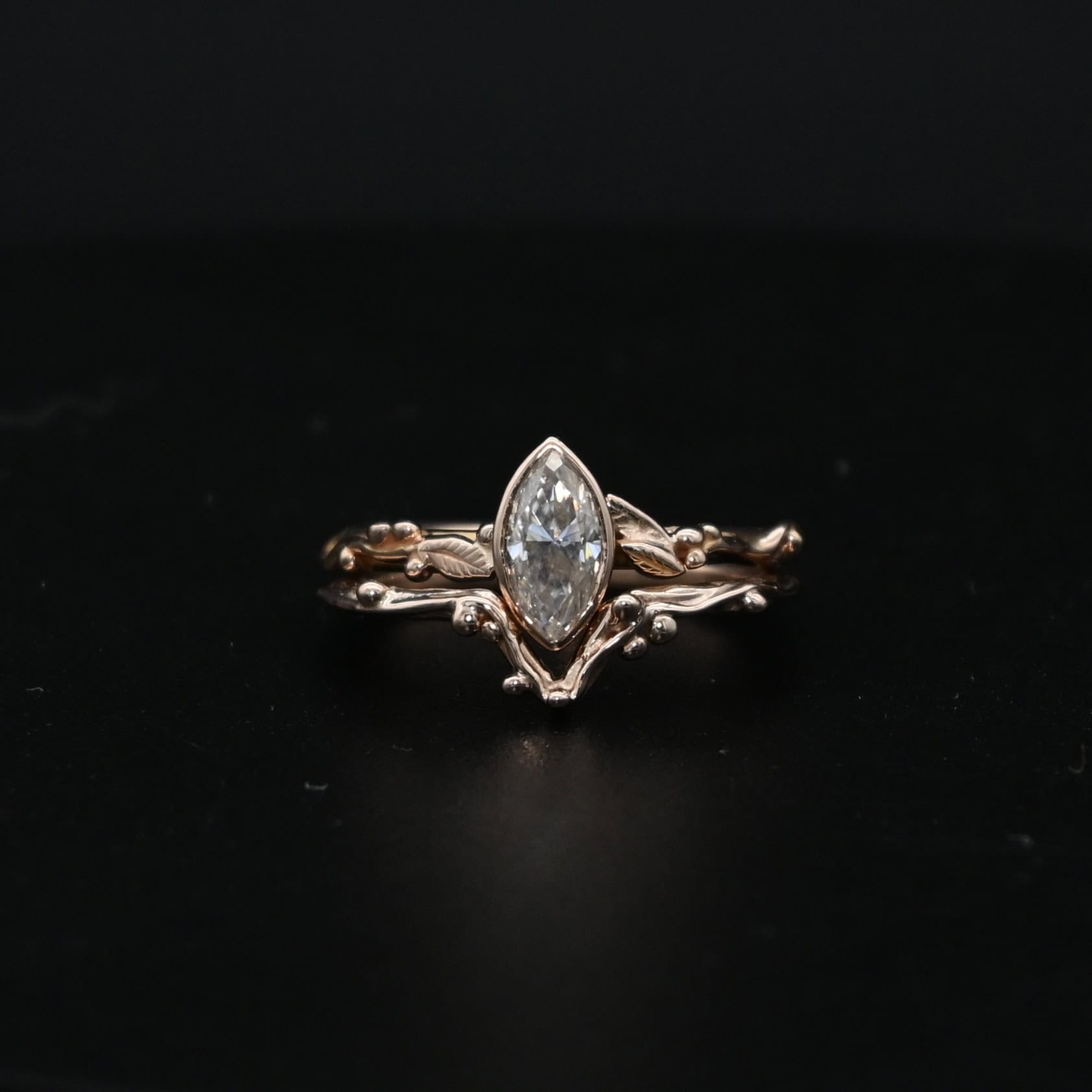 "Forrest Bride" Ring. There is a marquise shaped diamond in the center of the top band. The band is rose gold and has rose gold leaves and balls all around the band. The bottom band has a dip in the center to fit around the bottom of the marquise. This band has balls of rose gold around it as well.