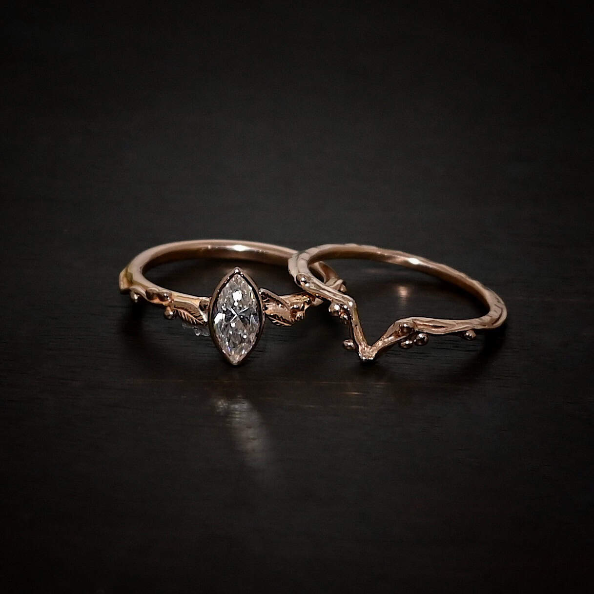 "Forrest Bride" Ring. There is a marquise shaped diamond in the center of the top band. The band is rose gold and has rose gold leaves and balls all around the band. The bottom band has a dip in the center to fit around the bottom of the marquise. This band has balls of rose gold around it as well.