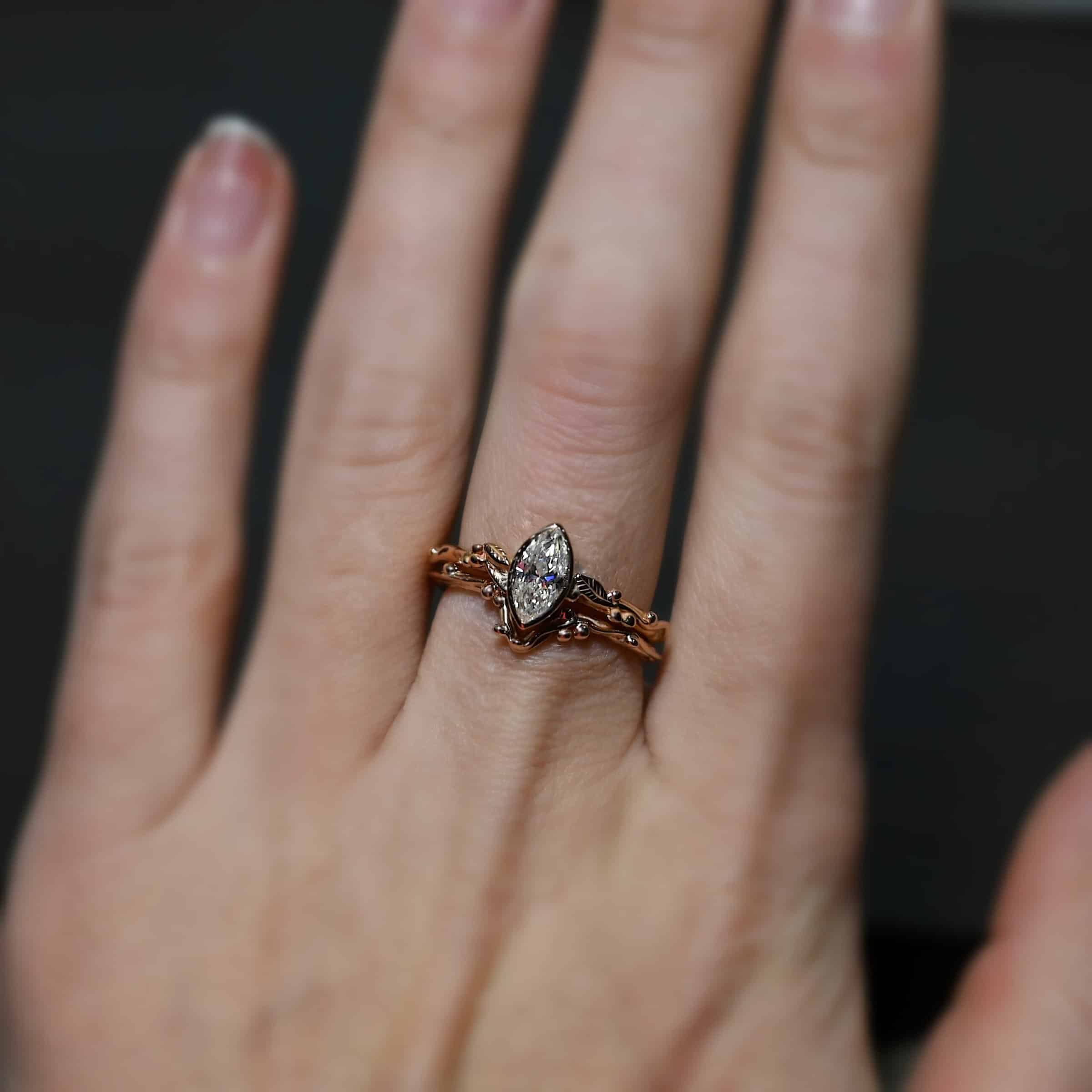 "Forrest Bride" Ring. There is a marquise shaped diamond in the center of the top band. The band is rose gold and has rose gold leaves and balls all around the band. The bottom band has a dip in the center to fit around the bottom of the marquise. This band has balls of rose gold around it as well. Ring is worn by a model.