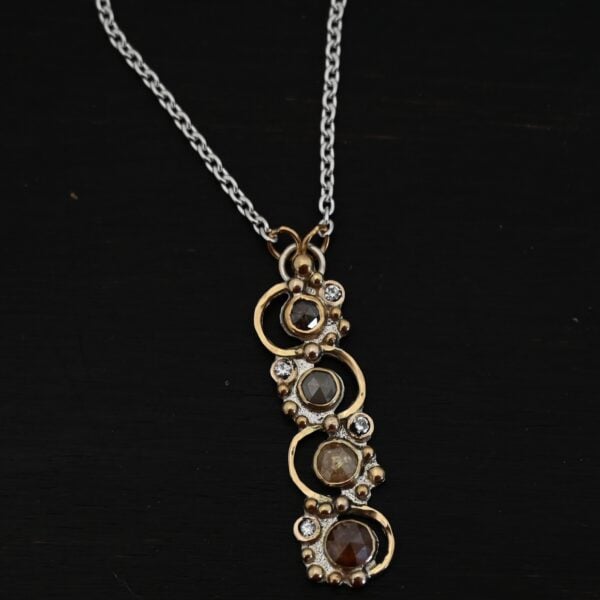 Photo of the "Music Of The Night" Necklace. Made out of sterling silver, gold, and diamonds. The top of the pendant, on the back, has a silver loop soldered to it with two gold jump rings that each end of the chain is attached to. There is gold ball detail that swirls back and forth all the way down the pendant. There is a line of hammered gold that also swirls around the pendant, but opposite of the gold balls, so they intertwine and intercept each other. There are eight diamonds on the pendant. Four of them are larger, and go in a straight line down the pendant. The other four are scattered around them. The gold line and gold balls twirl around each of the diamonds on opposite sides. Necklace is lying against a dark background.