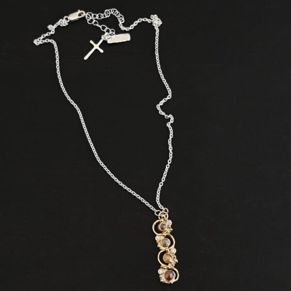Photo of the "Music Of The Night" Necklace. Made out of sterling silver, gold, and diamonds. The top of the pendant, on the back, has a silver loop soldered to it with two gold jump rings that each end of the chain is attached to. There is gold ball detail that swirls back and forth all the way down the pendant. There is a line of hammered gold that also swirls around the pendant, but opposite of the gold balls, so they intertwine and intercept each other. There are eight diamonds on the pendant. Four of them are larger, and go in a straight line down the pendant. The other four are scattered around them. The gold line and gold balls twirl around each of the diamonds on opposite sides. Necklace is lying against a dark background.