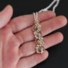 Photo of the "Music Of The Night" Necklace. Made out of sterling silver, gold, and diamonds. The top of the pendant, on the back, has a silver loop soldered to it with two gold jump rings that each end of the chain is attached to. There is gold ball detail that swirls back and forth all the way down the pendant. There is a line of hammered gold that also swirls around the pendant, but opposite of the gold balls, so they intertwine and intercept each other. There are eight diamonds on the pendant. Four of them are larger, and go in a straight line down the pendant. The other four are scattered around them. The gold line and gold balls twirl around each of the diamonds on opposite sides. Necklace is lying against the fingertips of a hand to demonstrate size.
