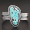 "Mountains Are Calling" Cuff. There is a large, oval shaped turquoise in the center with hammered sterling silver around the stone. The band of the cuff is thick and oxidized. The entire center of the cuff has a thick rectangular line that is cut out and oxidized deeper than the rest of the cuff.