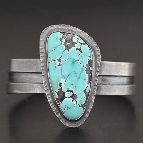 "Mountains Are Calling" Cuff. There is a large, oval shaped turquoise in the center with hammered sterling silver around the stone. The band of the cuff is thick and oxidized. The entire center of the cuff has a thick rectangular line that is cut out and oxidized deeper than the rest of the cuff.