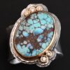 "Sunshine & Blue Skies" Ring. There is a large oval shaped turquoise, set slightly askew, pointing towards the right. The turquoise is set in gold, as well as the diamonds. There are three small diamonds on the top and two on the bottom. Both follow the curve of the oval. There are two layers of sterling silver around the stone, and both are hammered. Ring band is thick silver and hammered.