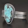 "Mountains Are Calling" Cuff. There is a large, oval shaped turquoise in the center with hammered sterling silver around the stone. The band of the cuff is thick and oxidized. The entire center of the cuff has a thick rectangular line that is cut out and oxidized deeper than the rest of the cuff.