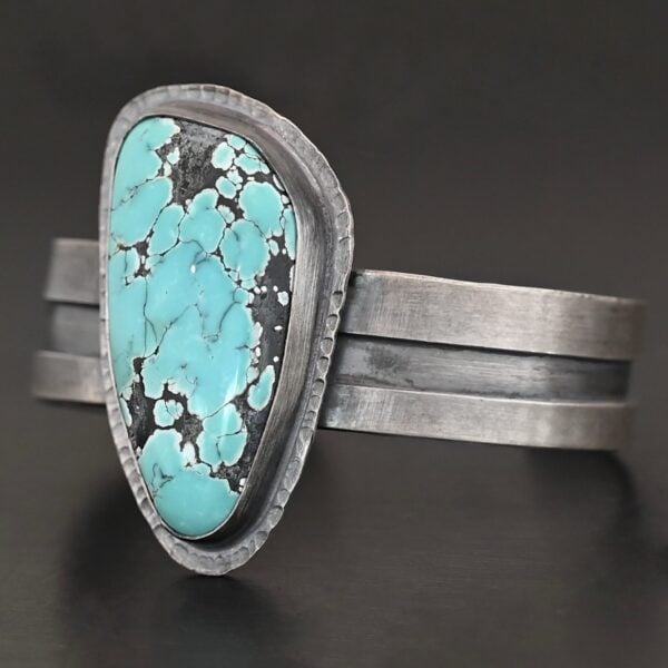 "Mountains Are Calling" Cuff. There is a large, oval shaped turquoise in the center with hammered sterling silver around the stone. The band of the cuff is thick and oxidized. The entire center of the cuff has a thick rectangular line that is cut out and oxidized deeper than the rest of the cuff.