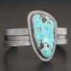 "Mountains Are Calling" Cuff. There is a large, oval shaped turquoise in the center with hammered sterling silver around the stone. The band of the cuff is thick and oxidized. The entire center of the cuff has a thick rectangular line that is cut out and oxidized deeper than the rest of the cuff.