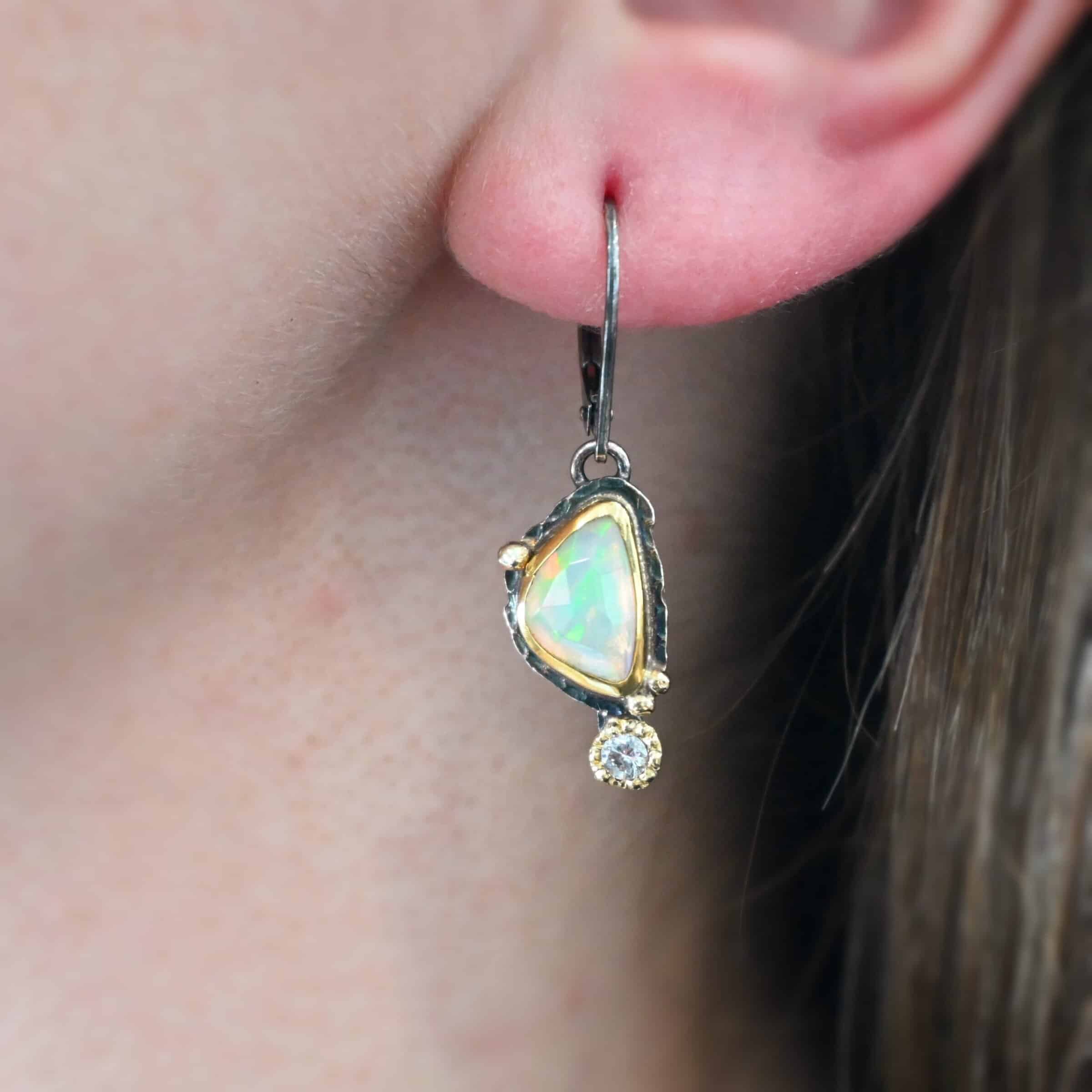 Light From Within Earrings The "Light From Within Earrings" are made out of sterling silver, gold, opal, and diamond. The opal is the center stone, with the diamond attached directly below the opal. There are three gold dots on the earring, and they are scattered around the center opal. The earring is worn by a model up close.