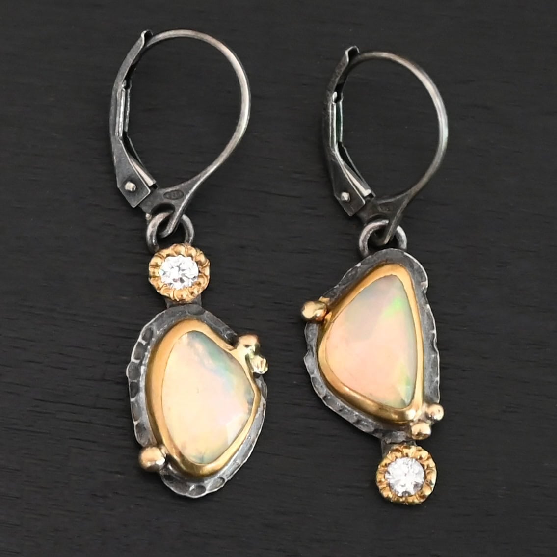 Light From Within Earrings New The "Light From Within Earrings" are made out of sterling silver, gold, opal, and diamond. The opal is the center stone, with the diamond attached directly above the opal on the left earring and below the opal on the right earring. There are three gold dots on the earrings, and they are scattered around the center opal. The earrings are lying against a dark background.