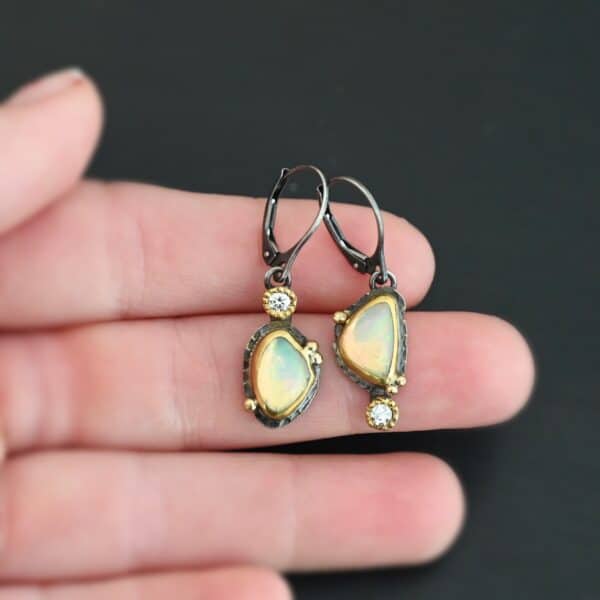 The "Light From Within Earrings" are made out of sterling silver, gold, opal, and diamond. The opal is the center stone, with the diamond attached directly above the opal on the left earring and below the opal on the right earring. There are three gold dots on the earrings, and they are scattered around the center opal. The earrings are held in the fingertips of a hand.