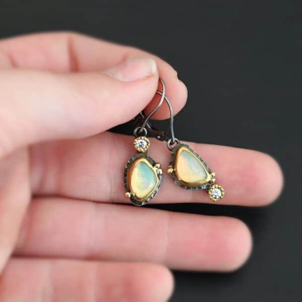 The "Light From Within Earrings" are made out of sterling silver, gold, opal, and diamond. The opal is the center stone, with the diamond attached directly above the opal on the left earring and below the opal on the right earring. There are three gold dots on the earrings, and they are scattered around the center opal. The earrings are held in the fingertips of a hand.