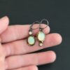 The "Color Of Moonrise Earrings" are made out of sterling silver, gold, opal, and diamond. The opal is the center stone, with the diamond attached directly above the opal. There are three gold dots on the earrings, and they are scattered around the center opal. The earrings are held in the fingertips of a hand.