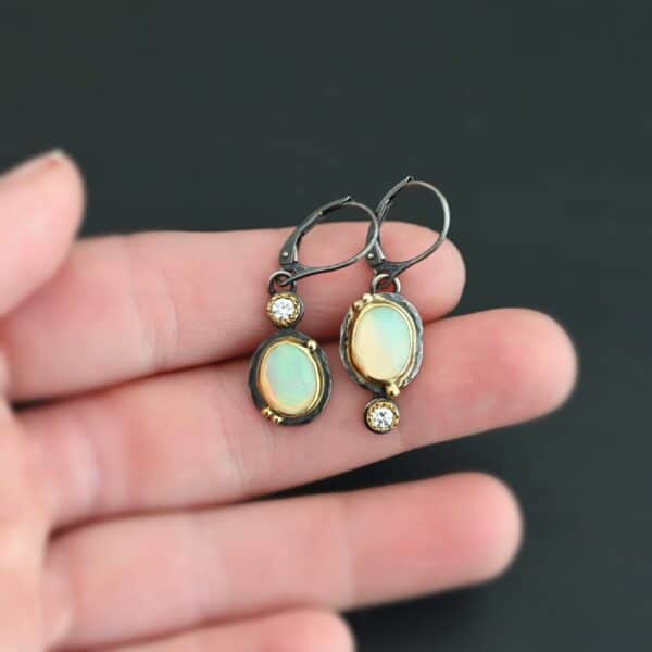 The "Color Of Moonrise Earrings" are made out of sterling silver, gold, opal, and diamond. The opal is the center stone, with the diamond attached directly above the opal. There are three gold dots on the earrings, and they are scattered around the center opal. The earrings are held in the fingertips of a hand.
