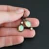 The "Color Of Moonrise Earrings" are made out of sterling silver, gold, opal, and diamond. The opal is the center stone, with the diamond attached directly above the opal. There are three gold dots on the earrings, and they are scattered around the center opal. The earrings are held in the fingertips of a hand.