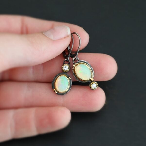 The "Color Of Moonrise Earrings" are made out of sterling silver, gold, opal, and diamond. The opal is the center stone, with the diamond attached directly above the opal. There are three gold dots on the earrings, and they are scattered around the center opal. The earrings are held in the fingertips of a hand.