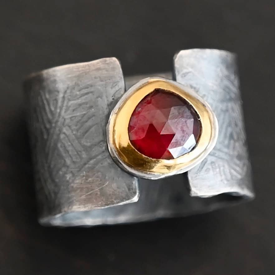 "Fire Of Love" Ring. Made out of sterling silver, gold, and garnet. Pear shaped garnet pointing up in the center of the ring set in gold. The gold sits on a layer of silver. The two sides of the band do not touch in the center. The garnet setting connects them together. The ring band is engraved with lines.