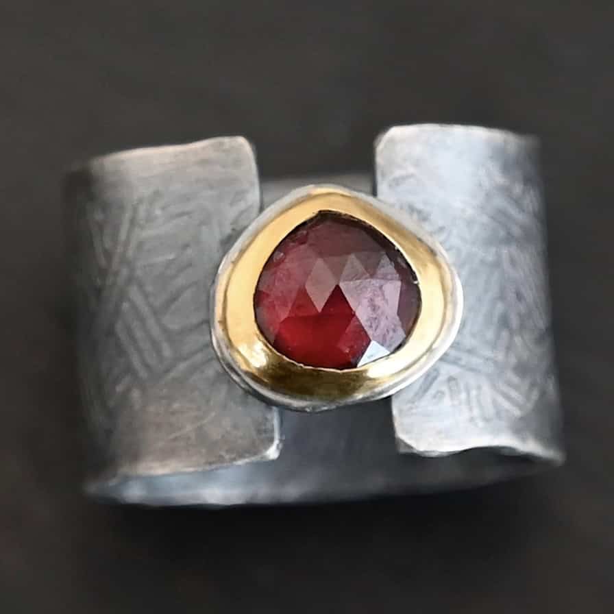 "Fire Of Love" Ring. Made out of sterling silver, gold, and garnet. Pear shaped garnet pointing up in the center of the ring set in gold. The gold sits on a layer of silver. The two sides of the band do not touch in the center. The garnet setting connects them together. The ring band is engraved with lines.