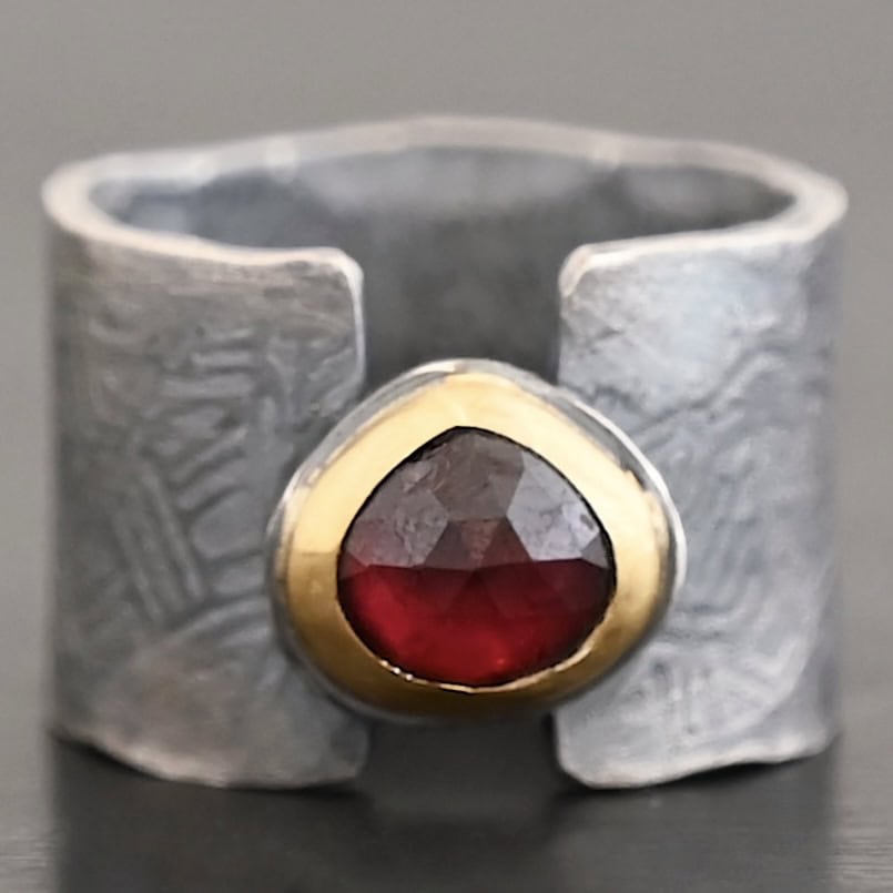 "Fire Of Love" Ring. Made out of sterling silver, gold, and garnet. Pear shaped garnet pointing up in the center of the ring set in gold. The gold sits on a layer of silver. The two sides of the band do not touch in the center. The garnet setting connects them together. The ring band is engraved with lines.