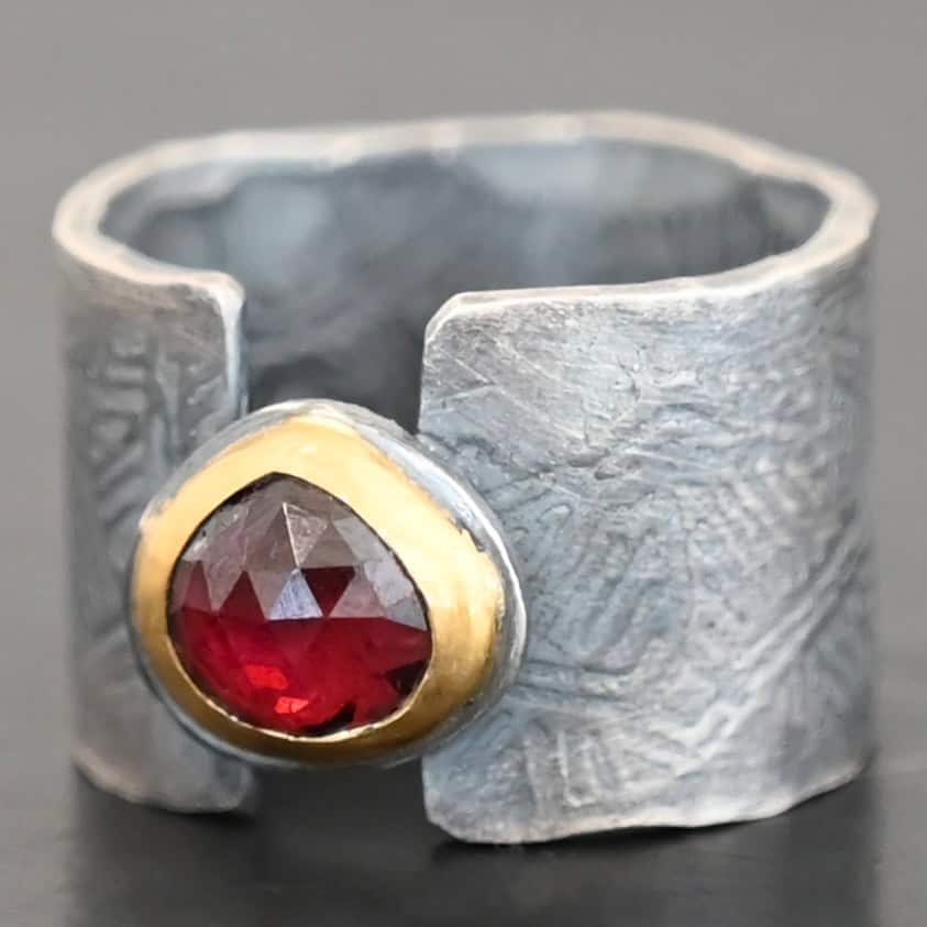 "Fire Of Love" Ring. Made out of sterling silver, gold, and garnet. Pear shaped garnet pointing up in the center of the ring set in gold. The gold sits on a layer of silver. The two sides of the band do not touch in the center. The garnet setting connects them together. The ring band is engraved with lines.